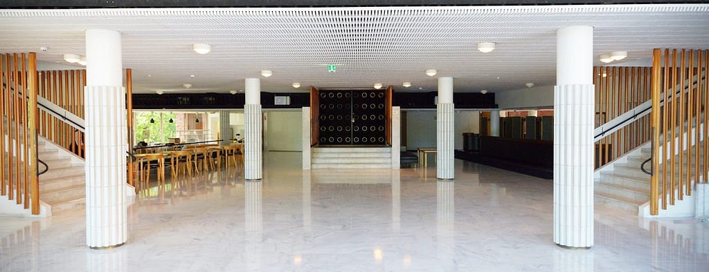 Jyväskylän yliopiston päärakennuksen aula. Kuva: Inna Lisko. Kuvassa näkyy vaaleaa marmorilattiaa, valkoisia koristeellisia pylväitä ja keskellä yliopiston juhlasalin pääovet. Portaikkojen lähtöpisteet näkyvät oikealla ja vasemmalla, kahvion tuoleja ja ikkuna vehreään ulkoilmaan näkyy vasemmalla sekä naulakkoja oikealla.